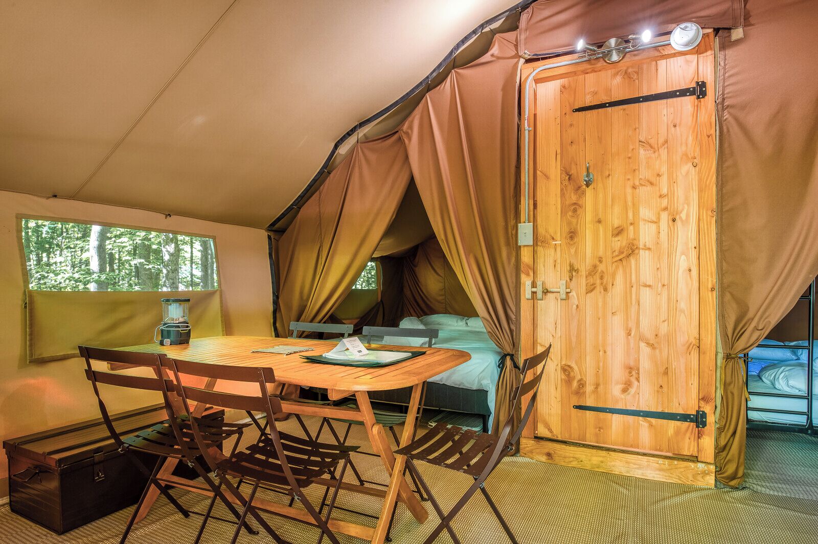 A cozy tented room with a wooden table, chairs, a bed, and a window overlooking a forest.