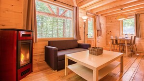 A cozy cabin interior with a wood stove, a sofa, a dining table, and large windows offering a view of the forest. - Huttopia White Mountains (Conway)