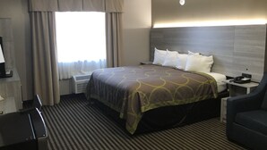 A hotel room with a bed, a desk, a chair, a nightstand, a lamp, and a window with curtains. - DuPont Suites (Louisville)