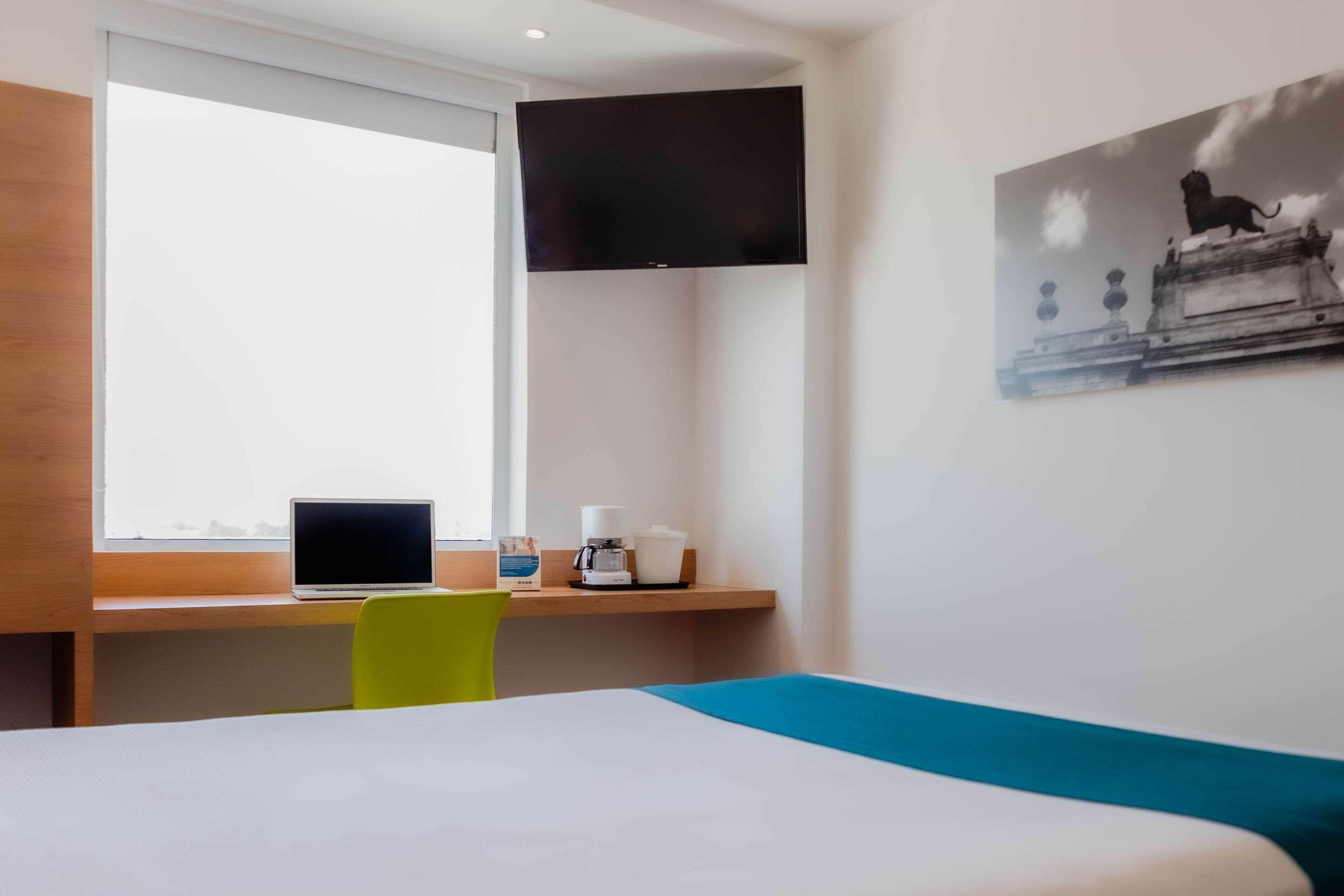 A hotel room with a bed, a desk with a laptop, a coffee maker, and a television mounted on the wall.
