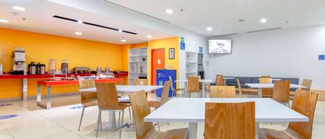A modern cafeteria with white tables and chairs, a counter with food items, and a television mounted on the wall.