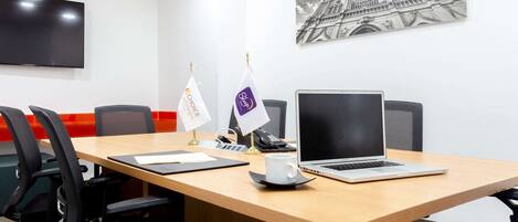 Een vergaderzaal met een houten tafel, zwarte stoelen, een laptop, een telefoon en twee vlaggen.