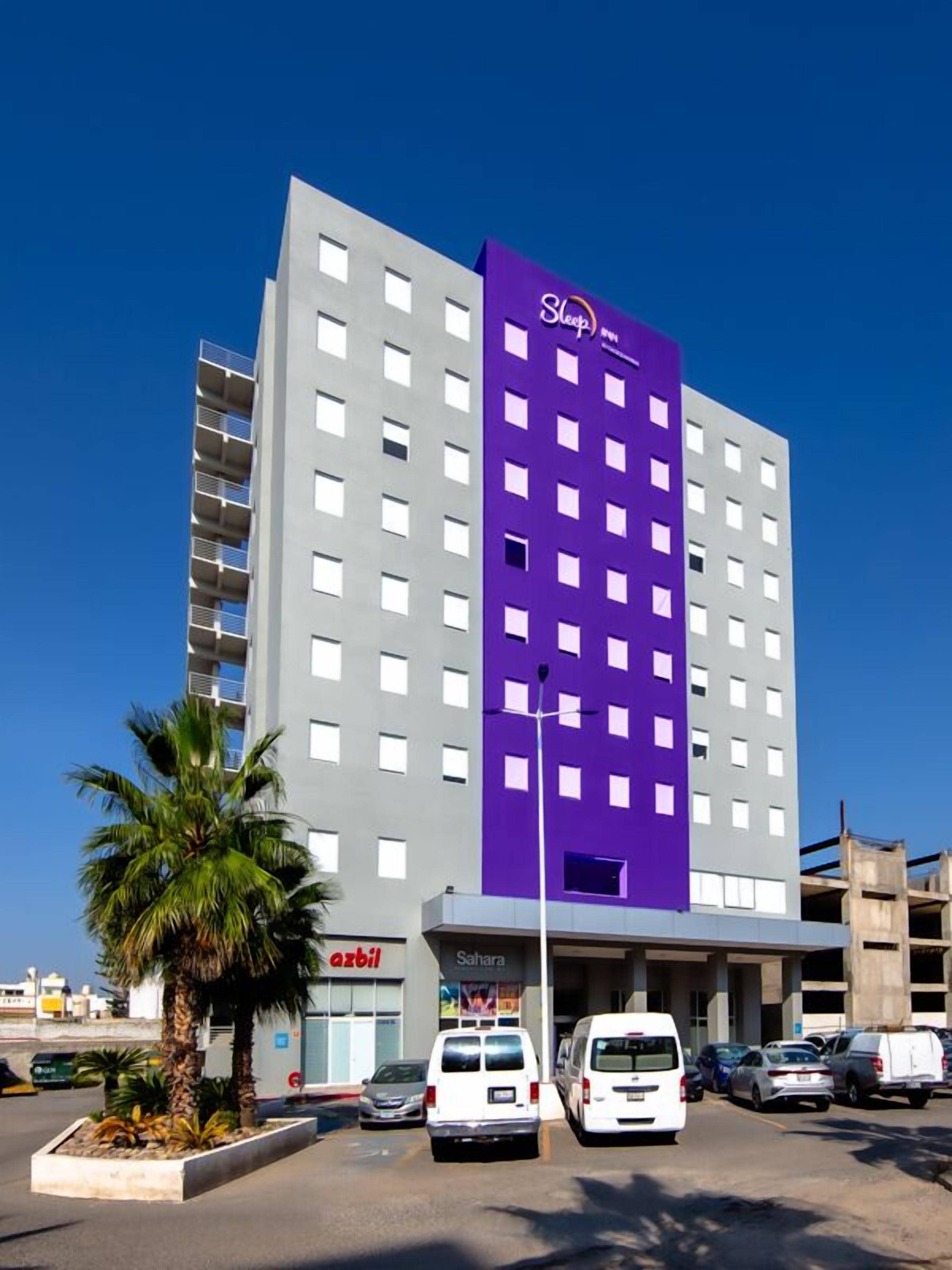 A multi-story hotel with a purple and grey facade, a parking lot with several white vans, and a palm tree in the foreground.