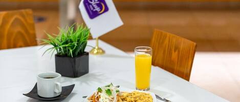 Een eettafel met een bord eten, een glas sinaasappelsap, een kop koffie en een kleine plant.