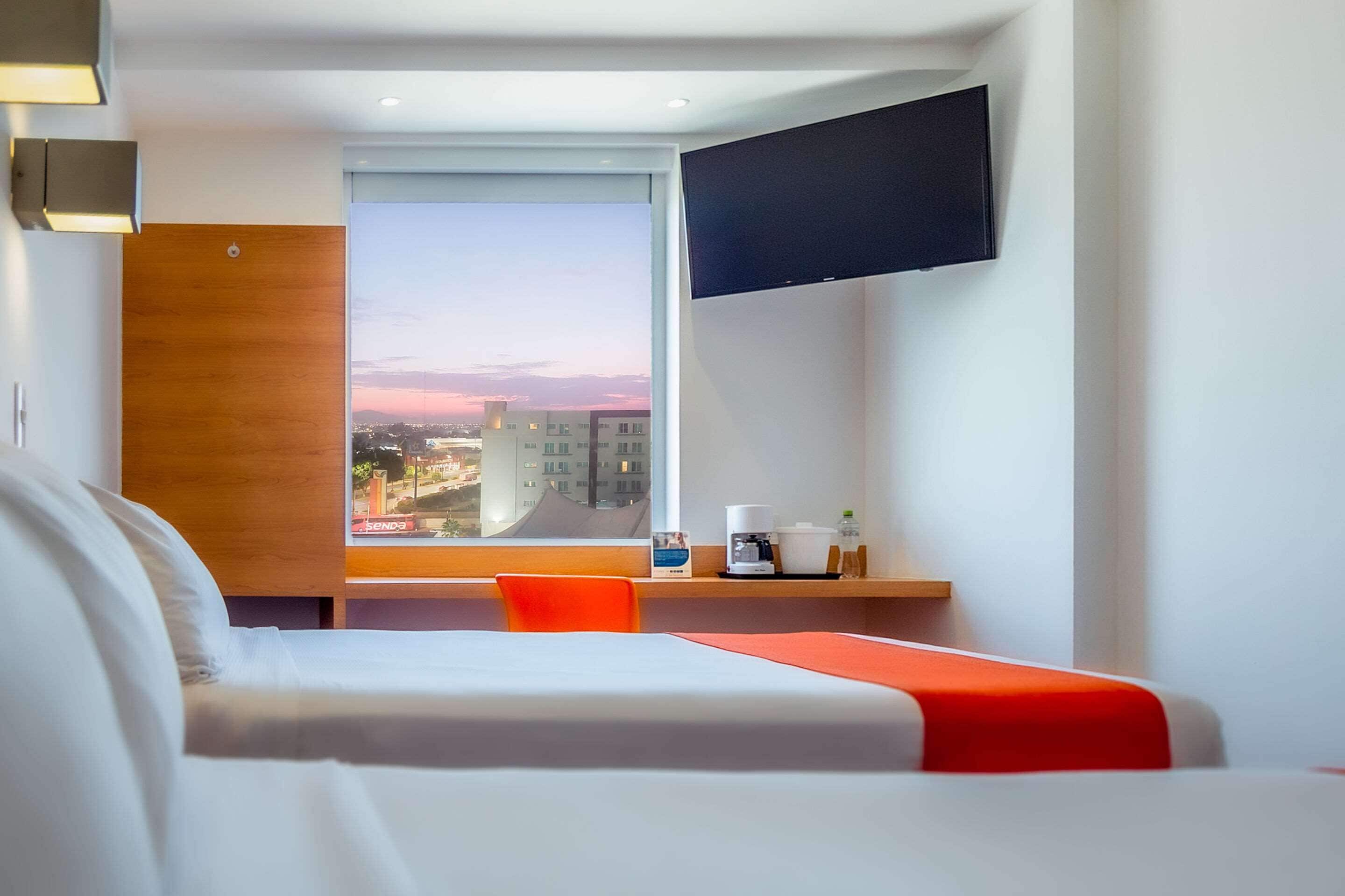 Una habitación de hotel con una cama, un escritorio con cafetera, un televisor montado en la pared y una ventana con vista a la ciudad.