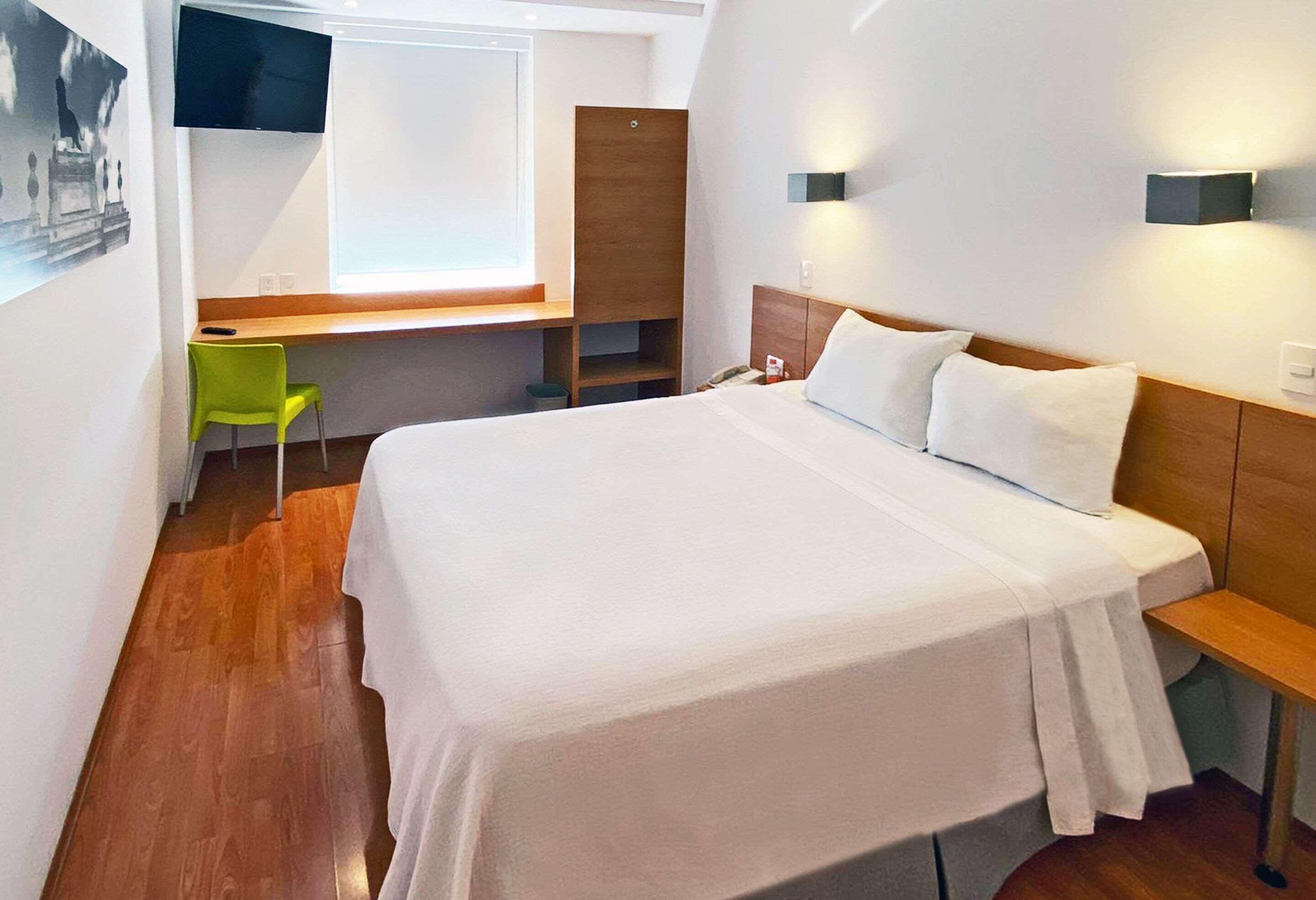 Une chambre d’hôtel équipée d’un lit, d’un bureau, d’une chaise, d’une télévision et d’une lampe fixée au mur.
