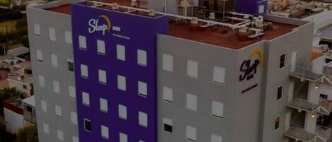 Un hotel de varios pisos con fachada púrpura y blanca, situado en una zona urbana con un atardecer de fondo.