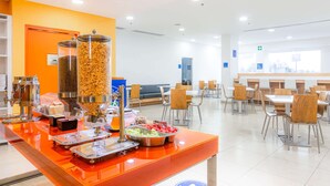 Una cafetería moderna y luminosa con una larga barra repleta de dispensadores y bandejas, rodeada de mesas y sillas.