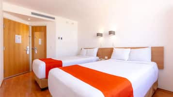 A hotel room with two beds, a wooden floor, and a white wall with two wall-mounted lights.