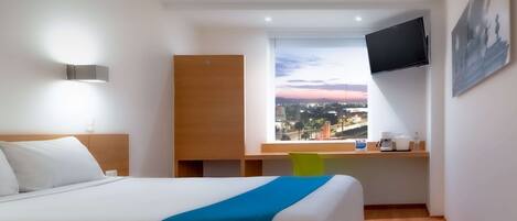 Una habitación de hotel con una cama, un escritorio con silla, un televisor montado en la pared y un ventanal con vista a la ciudad.