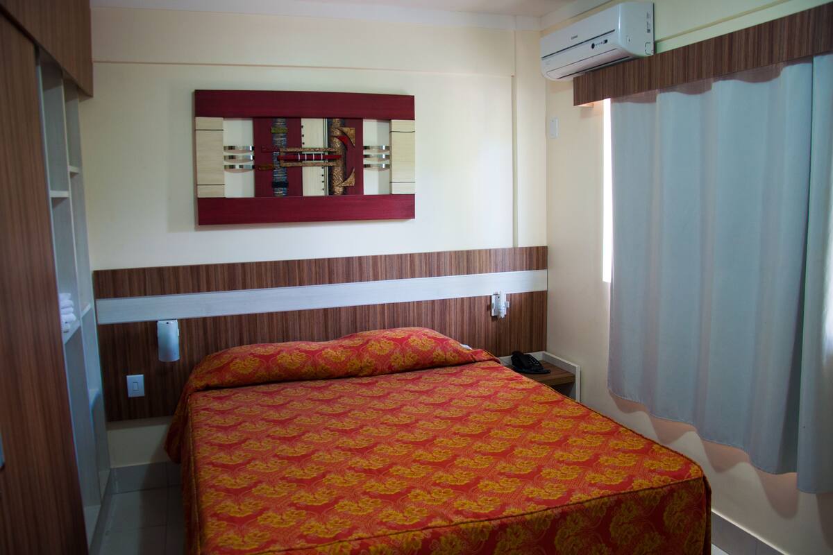 A hotel room with a bed, a mirror, an air conditioning unit, and a curtain.