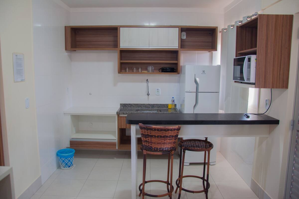 A compact kitchen with a refrigerator, microwave, and wooden cabinets. There are two wicker bar stools and a blue trash bin.