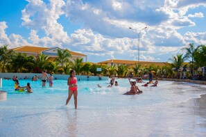 A swimming pool area with people enjoying the water, palm trees, and buildings in the background. - Apartment ABD Turismo - Lacqua DiRoma (Caldas Novas)