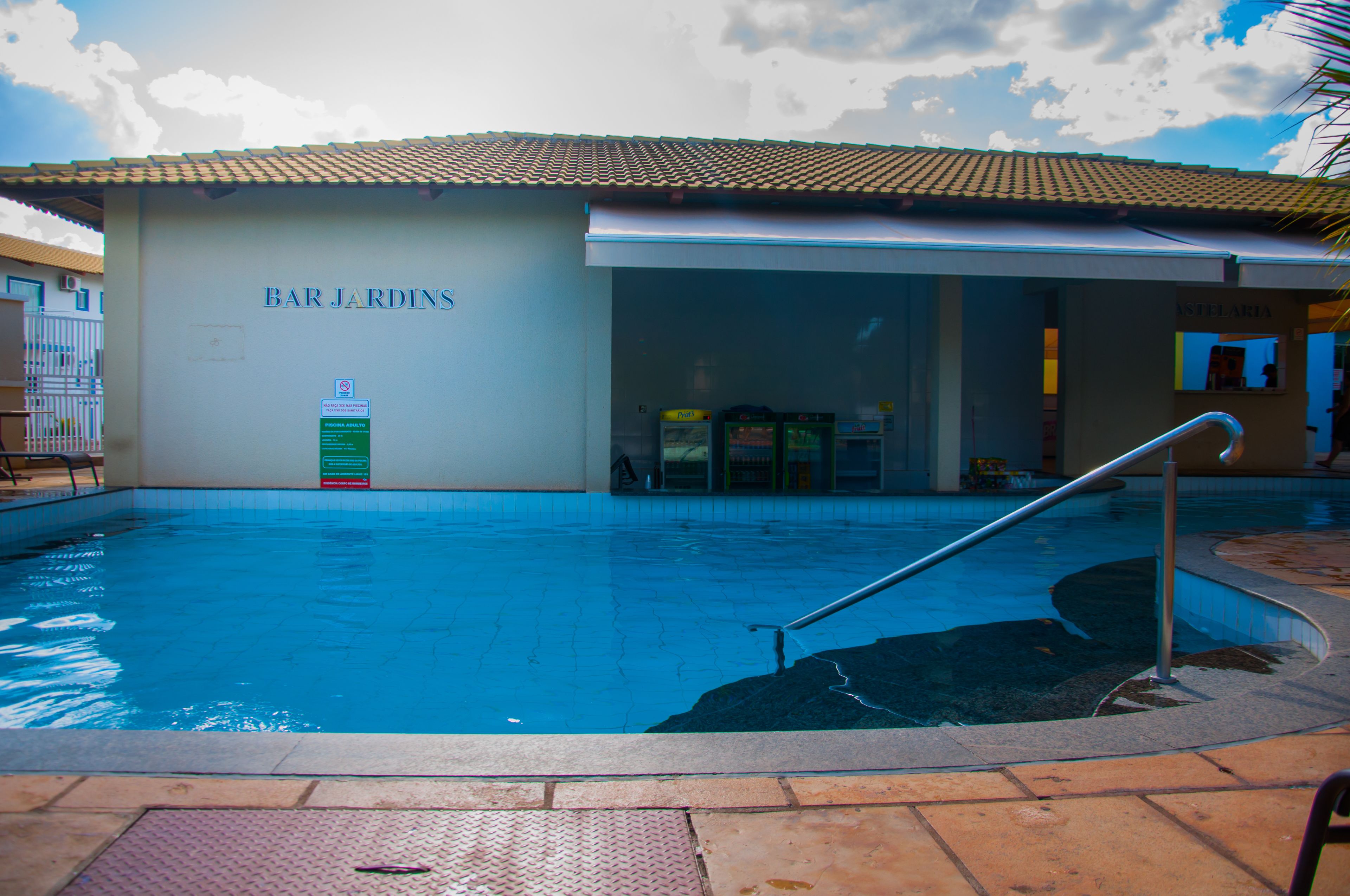Un espace piscine avec un bâtiment portant l’inscription « BAR JARDINS » et un panneau indiquant la présence d’un restaurant.