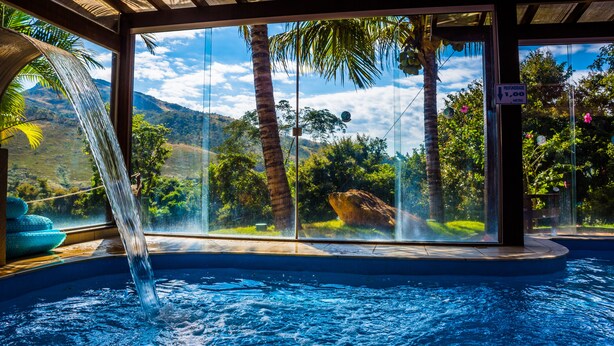 Un jacuzzi con caída de agua, rodeado por paredes de vidrio que ofrecen vistas a palmeras y montañas.