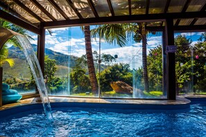A hot tub with a waterfall feature, surrounded by glass walls offering a view of palm trees and mountains. - Balneário do Lago Hotel (Capitólio)
