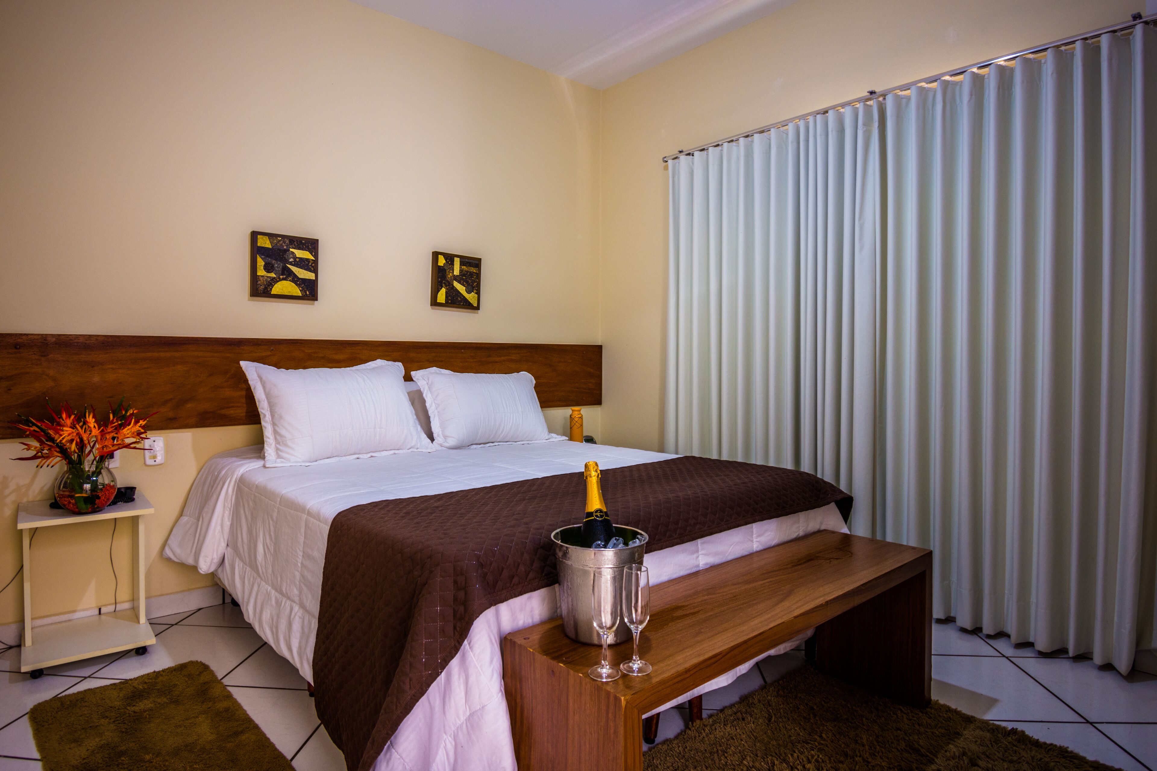 Een hotelkamer met een bed, een nachtkastje met een fles champagne, een houten bankje en twee decoratieve wandpanelen.