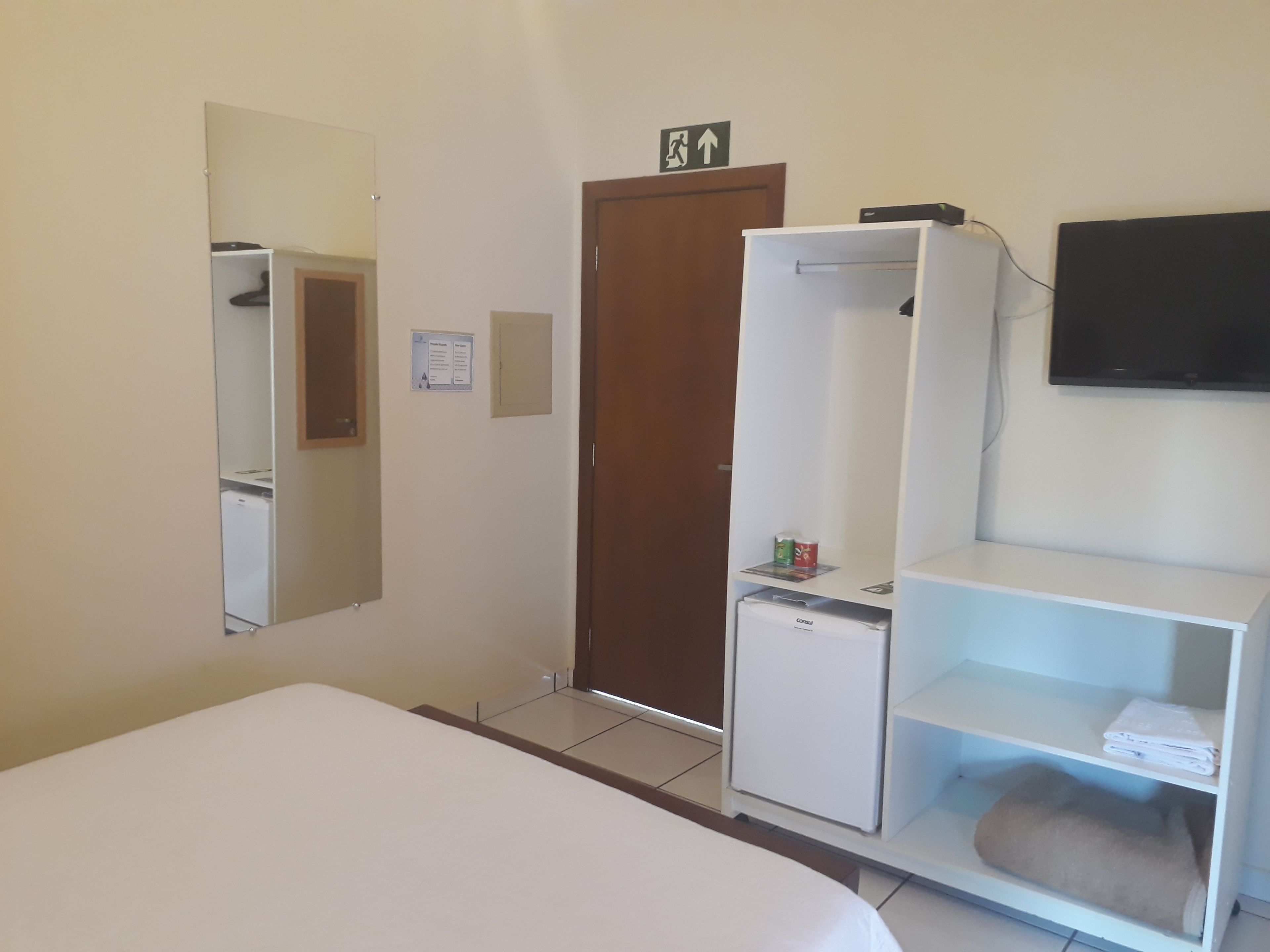 Habitació d'hotel amb un llit, un armari, un petit frigorífic, un mirall, una televisió i una porta amb el senyal d'eixida d'emergència.