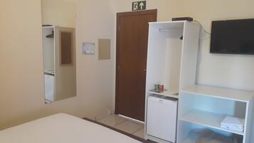 Habitació d'hotel amb un llit, un armari, un petit frigorífic, un mirall, una televisió i una porta amb el senyal d'eixida d'emergència.