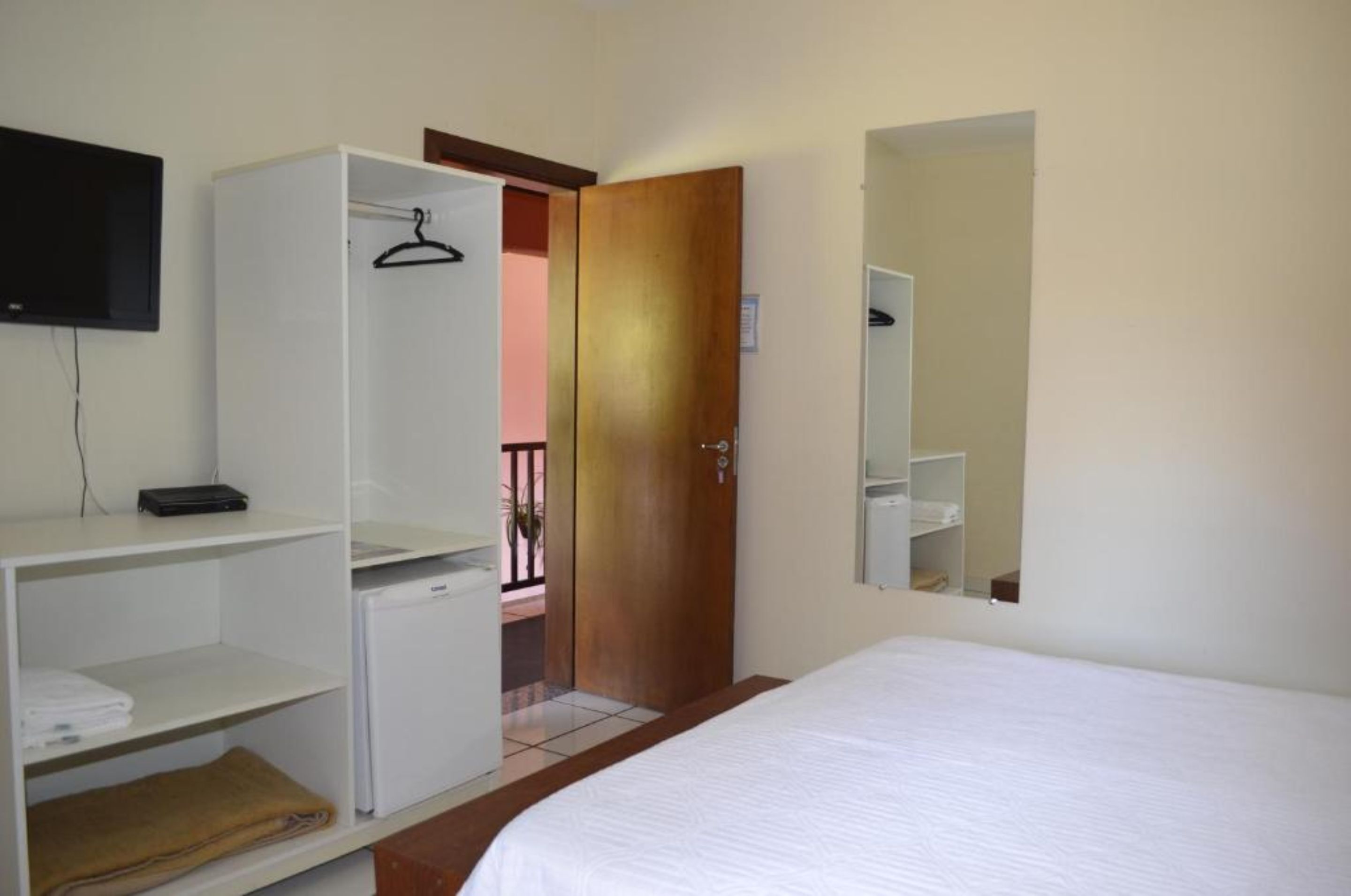 A hotel room with a bed, a wardrobe, a small desk with a computer, a TV mounted on the wall, and a door leading to a balcony.