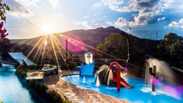 Una piscina con tobogán, fuentes de agua y montañas al fondo.