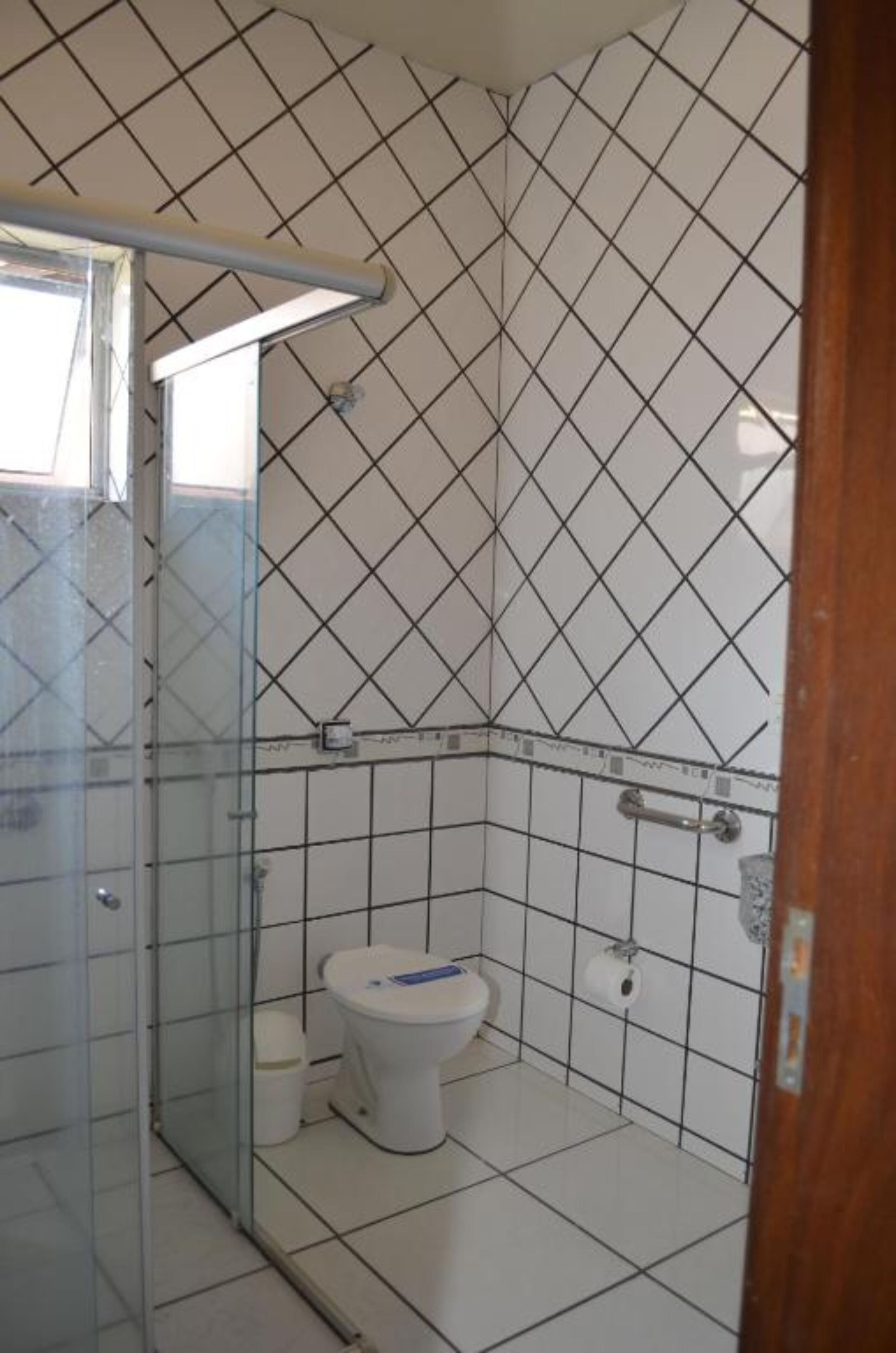 Un baño con suelo y paredes de azulejos blancos, cabina de ducha de vidrio, inodoro blanco y pasamanos visible.