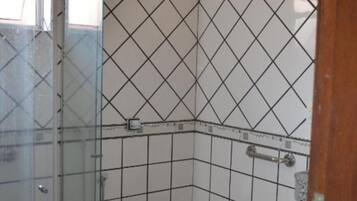 Un baño con suelo y paredes de azulejos blancos, cabina de ducha de vidrio, inodoro blanco y pasamanos visible.