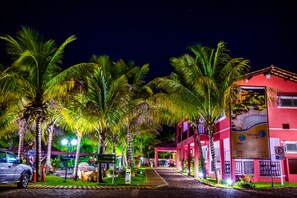 Vista nocturna de una calle bordeada de palmeras y un edificio iluminado con un cartel que indica recepción.