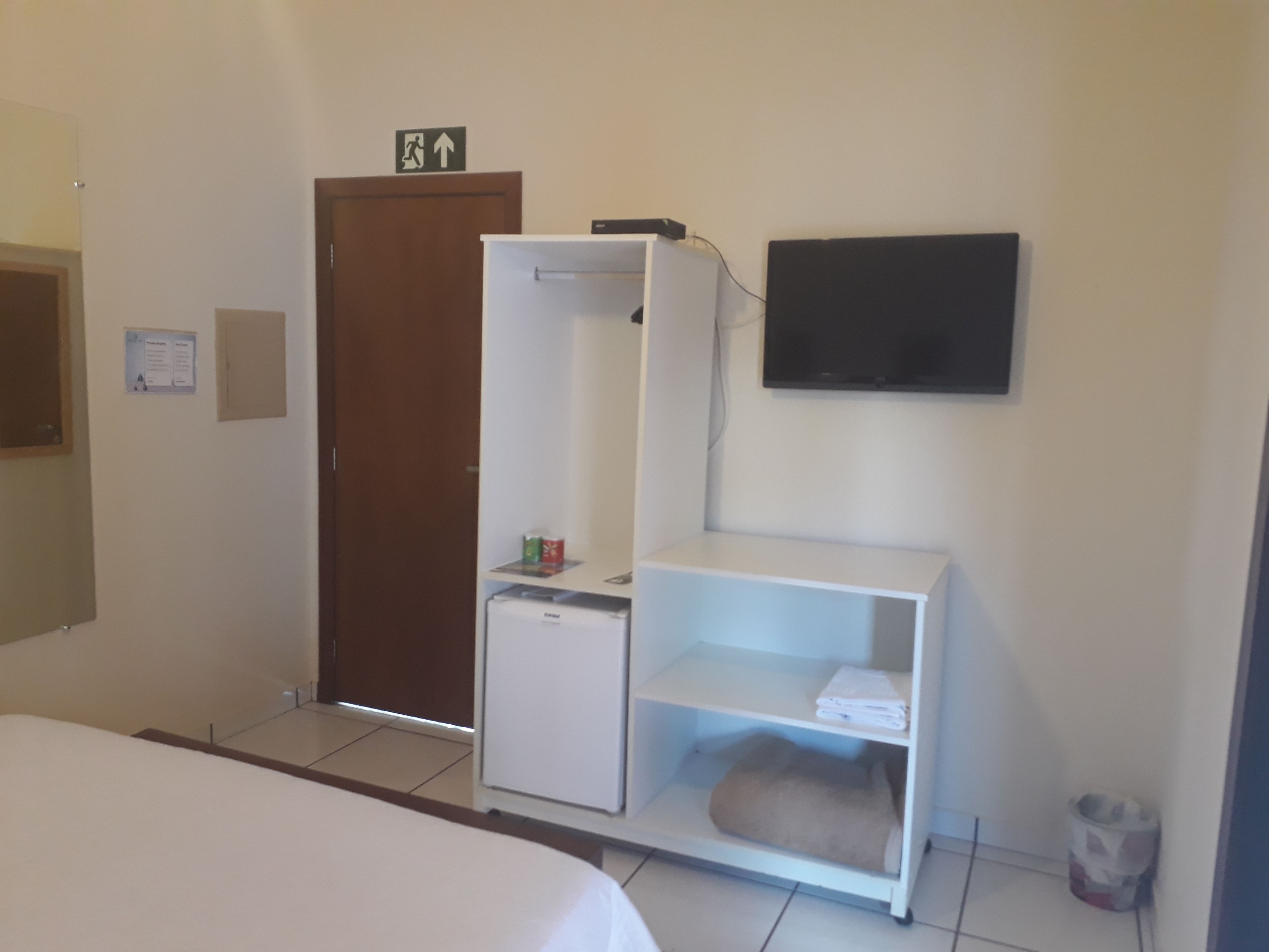 Habitación de hotel con cama, armario, refrigerador pequeño, estante con toallas, cubo de basura y televisor en la pared.