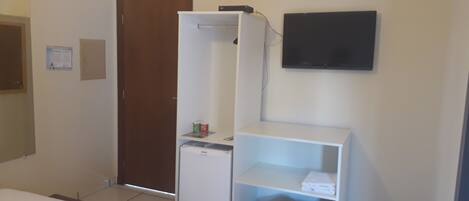 Habitación de hotel con cama, armario, refrigerador pequeño, estante con toallas, cubo de basura y televisor en la pared.