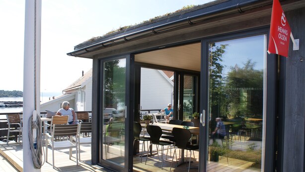 An outdoor dining area with wooden tables and chairs, a glass door entrance, and a view of the sea. - Fjordbris Hotell (Stavanger)
