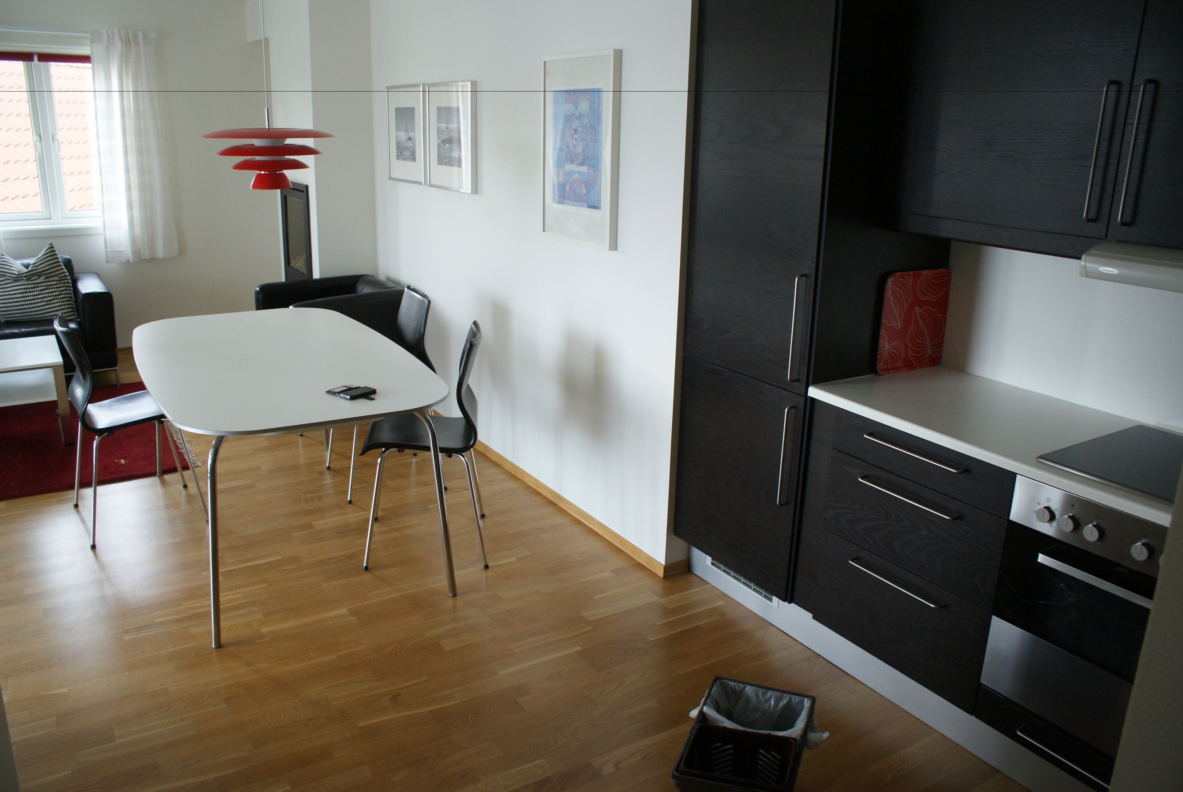 Una cocina moderna con armarios oscuros, encimera blanca y horno empotrado. Hay una zona de comedor con una mesa y sillas blancas, y una lámpara colgante roja.