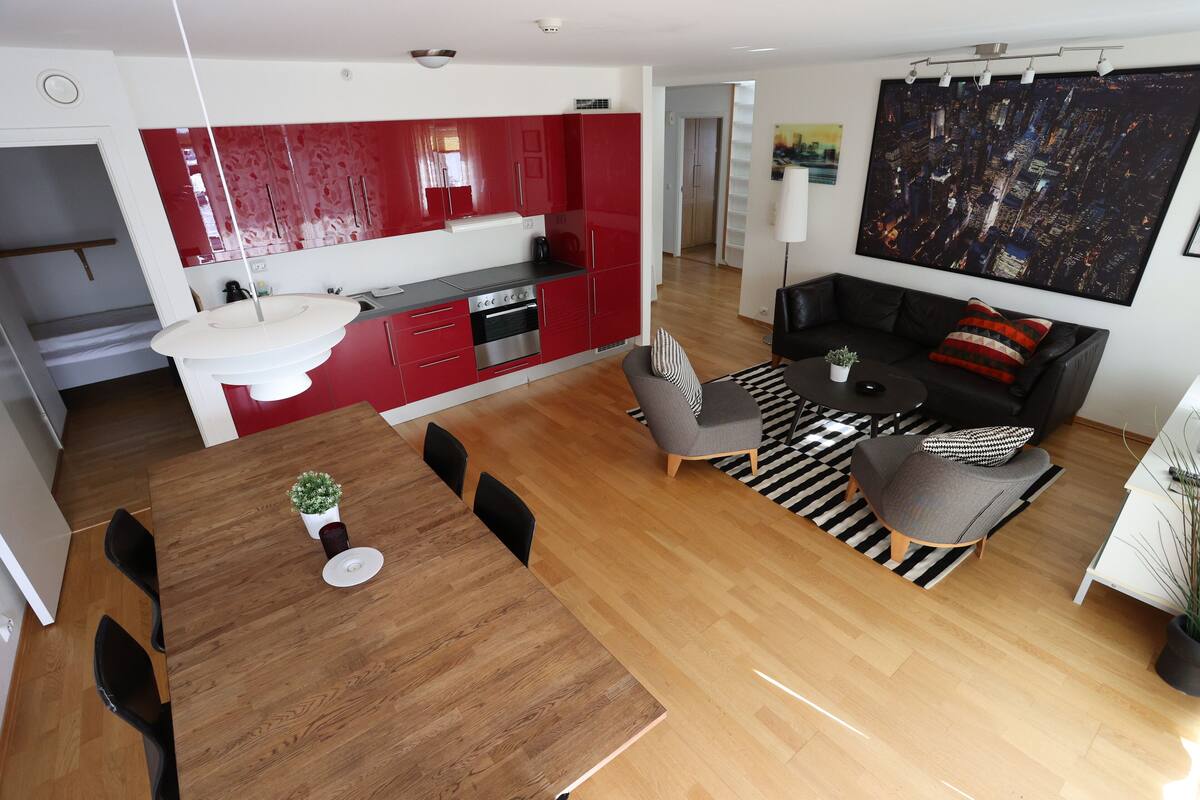 Eine moderne Küche mit roten Schränken, ein Essbereich mit Holztisch und -stühlen sowie ein Wohnzimmer mit einer Couch und einem Stadtbild.