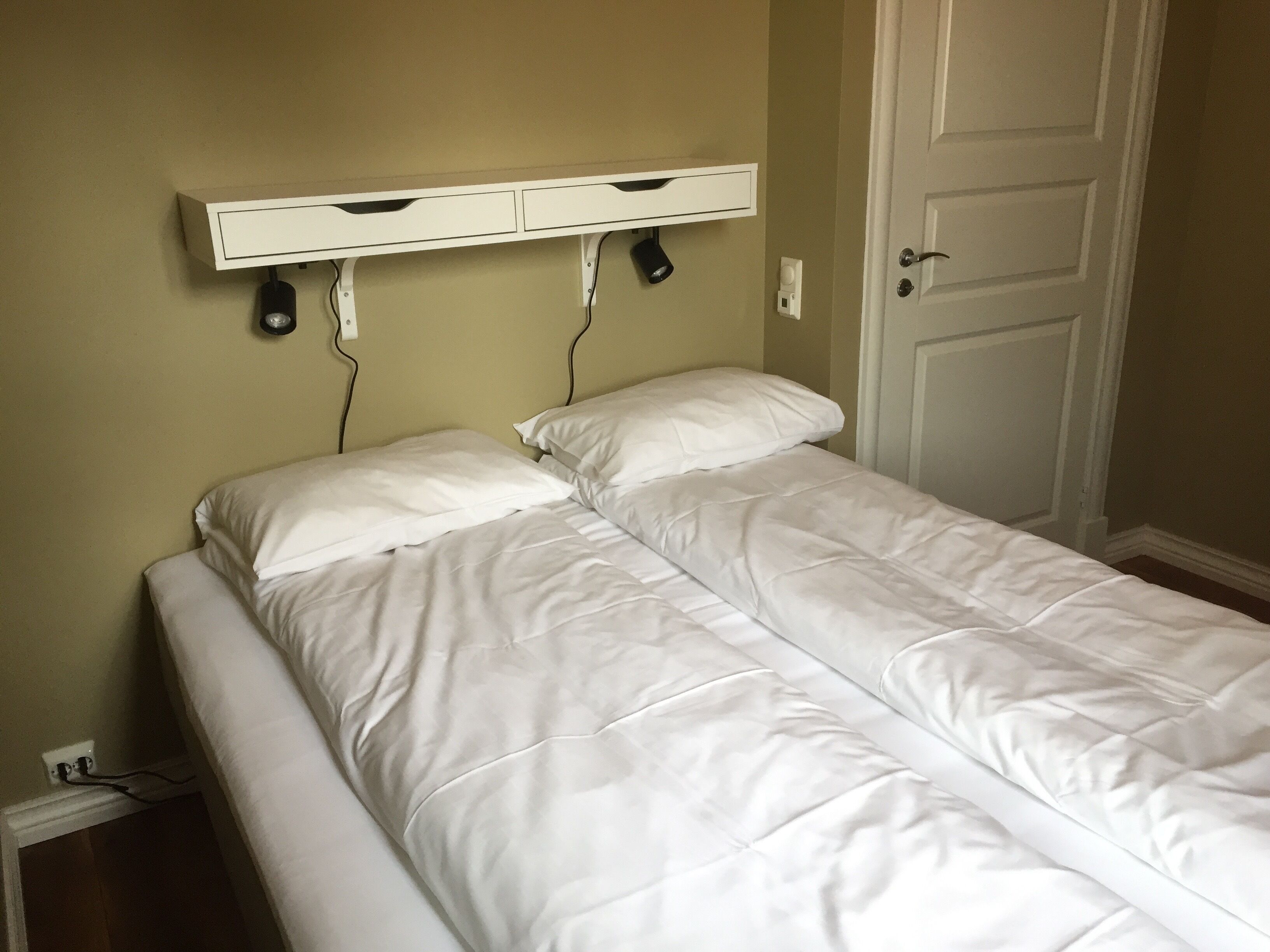 A single bed with white bedding, a bedside table with drawers, and a wall-mounted light fixture.