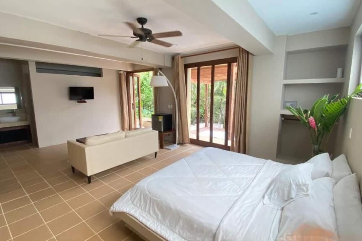 Un dormitorio con una cama grande, ventilador de techo, televisión, baño con lavamanos y vistas a zonas verdes.