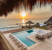 Zona de piscina con sillones y sombrillas, con vista al océano al atardecer.