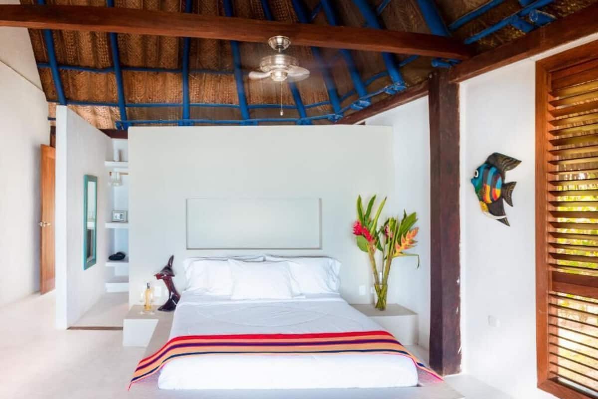 Un dormitorio con cama, mesitas de noche, lámpara de noche, ventilador de techo, ventana con persianas y un pez decorativo en la pared.