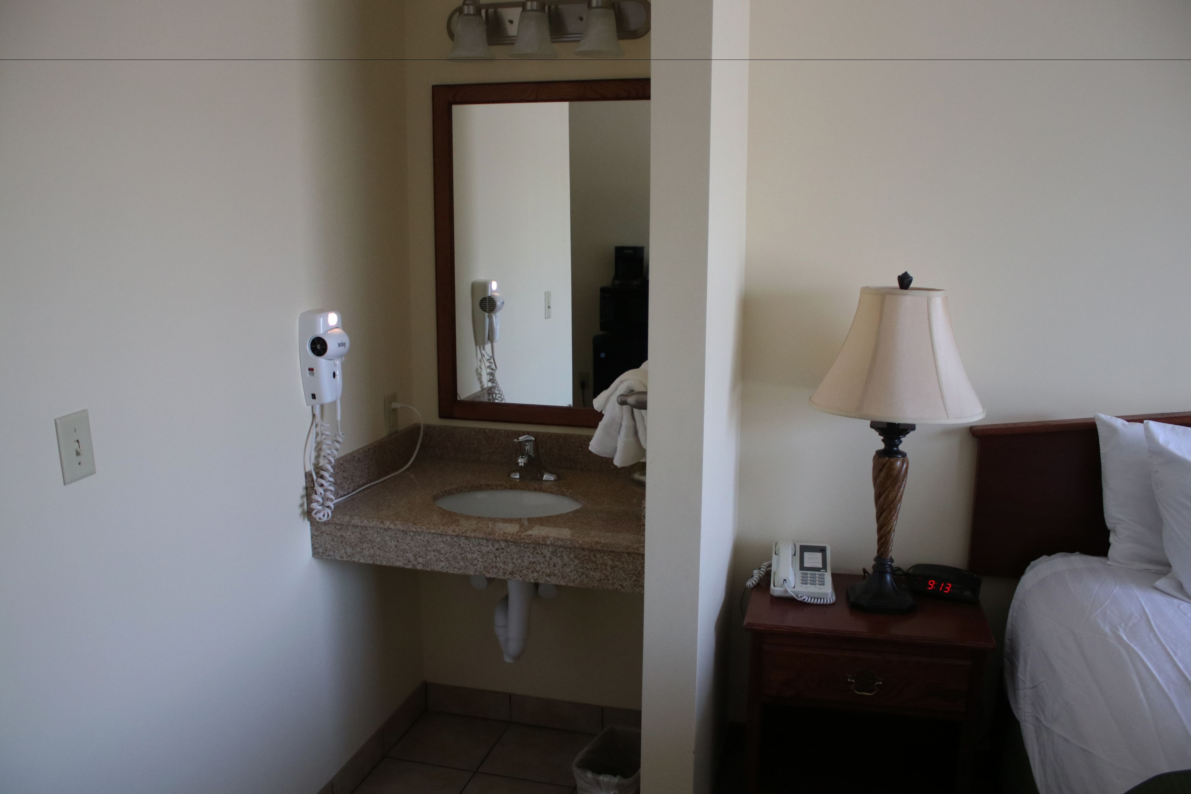 Un baño de hotel con lavamanos, espejo, secador de pelo y una mesita de noche con lámpara y reloj.