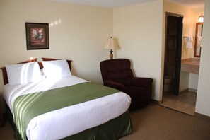 A hotel room with a bed, a chair, a lamp, and a bathroom visible through an open door. - Edgewood Hotel & Suites (Fairbury)