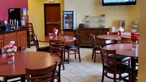 A restaurant with wooden tables and chairs, a refrigerator, and a television displaying a weather forecast. - Edgewood Hotel & Suites (Fairbury)