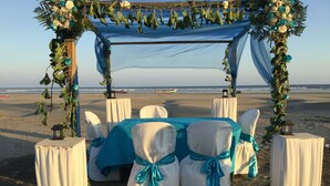 Een bruiloftdecoratie op het strand, met een luifel, witte stoelen met turquoise linten, en een tafel bedekt met een blauwe doek.