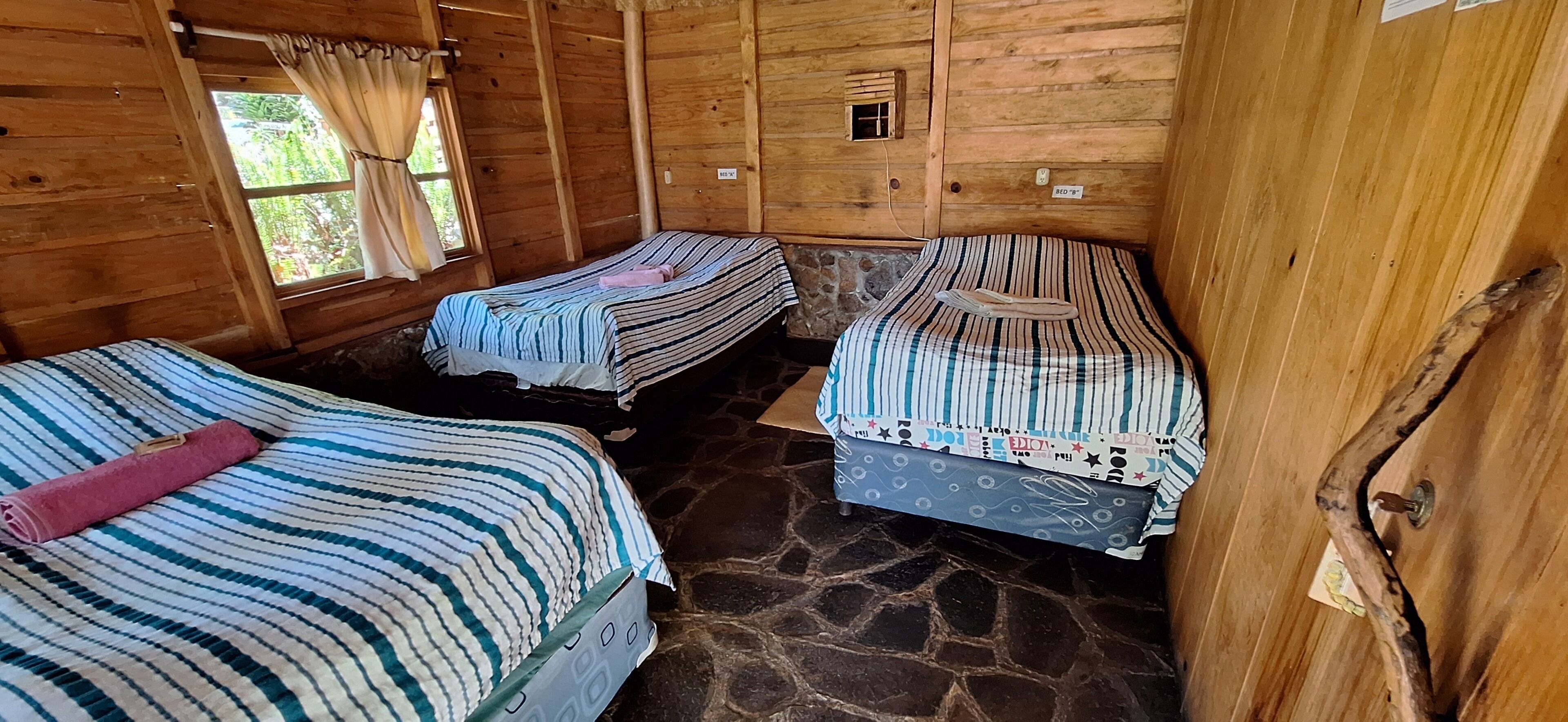A wooden cabin with two beds, a stone floor, and a window with curtains.
