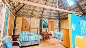 Ein rustikaler Raum mit Strohdach, Holzböden und schlichter Einrichtung, darunter ein Bett, Stühle und ein Tisch.