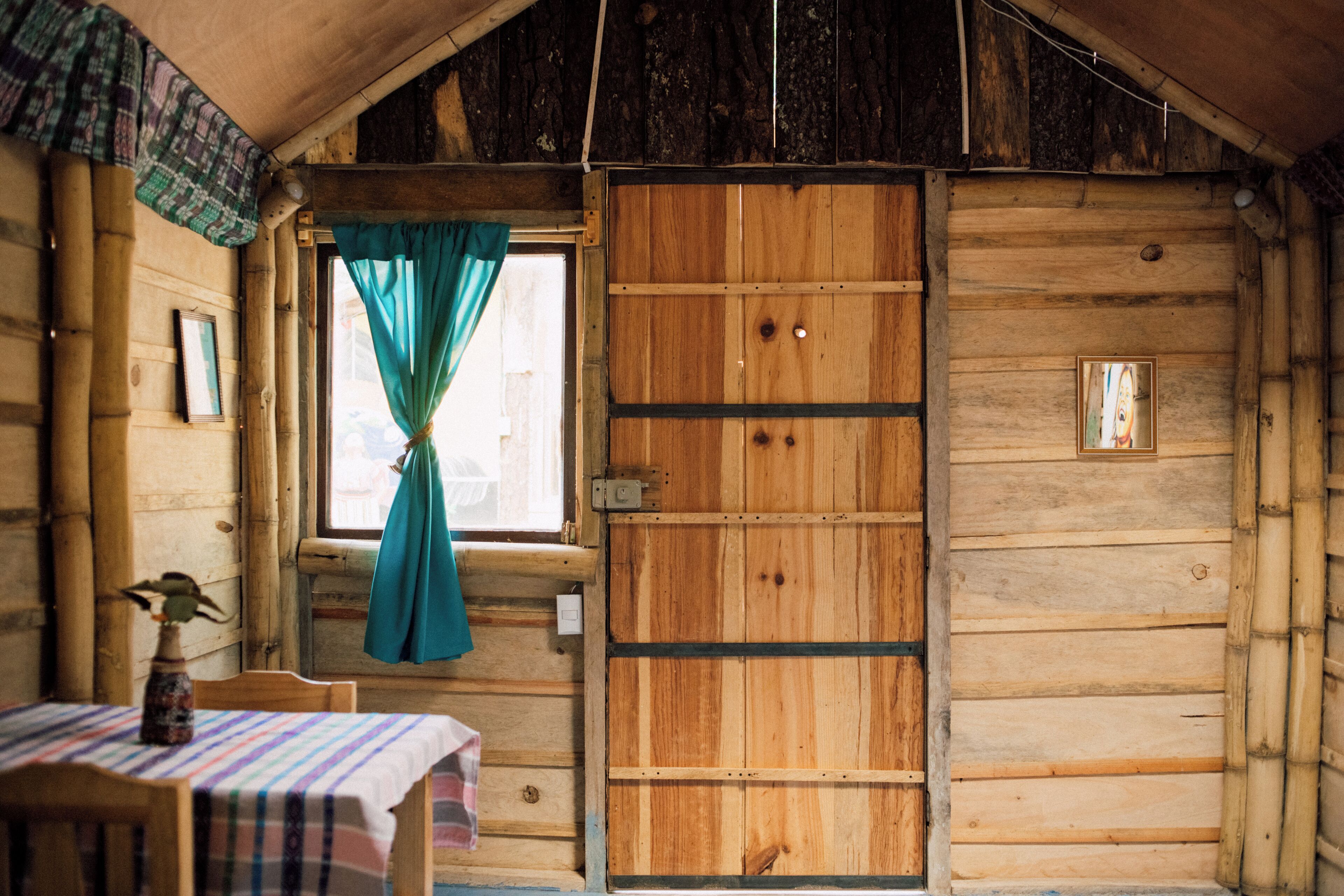 A wooden cabin interior with a table, chairs, a window with curtains, and a door.