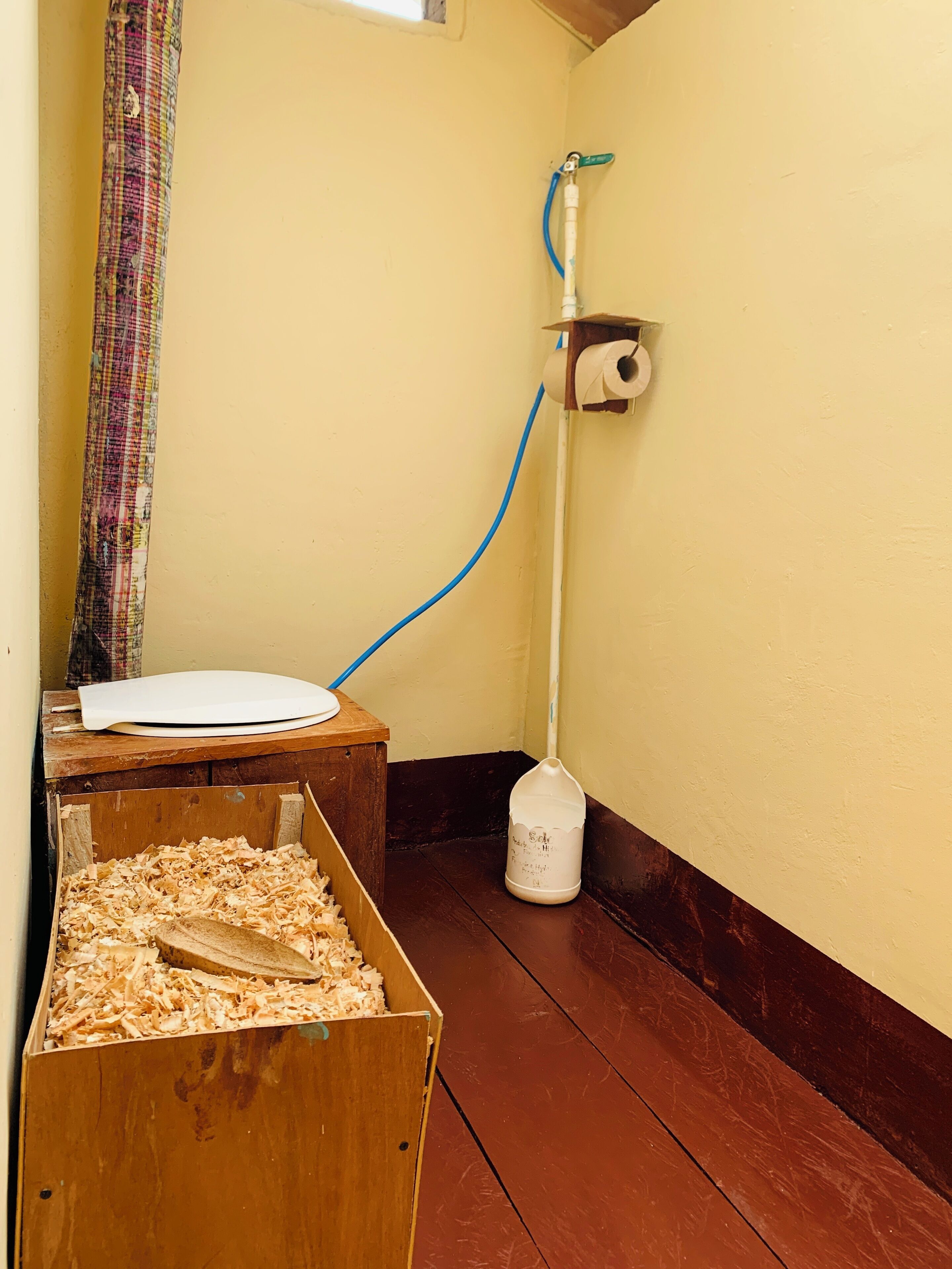 Un racó d'una habitació amb una caixa de fusta plena de virutes, un suport de paper higiènic blanc a la paret i una mànega blava connectada a una font d'aigua.