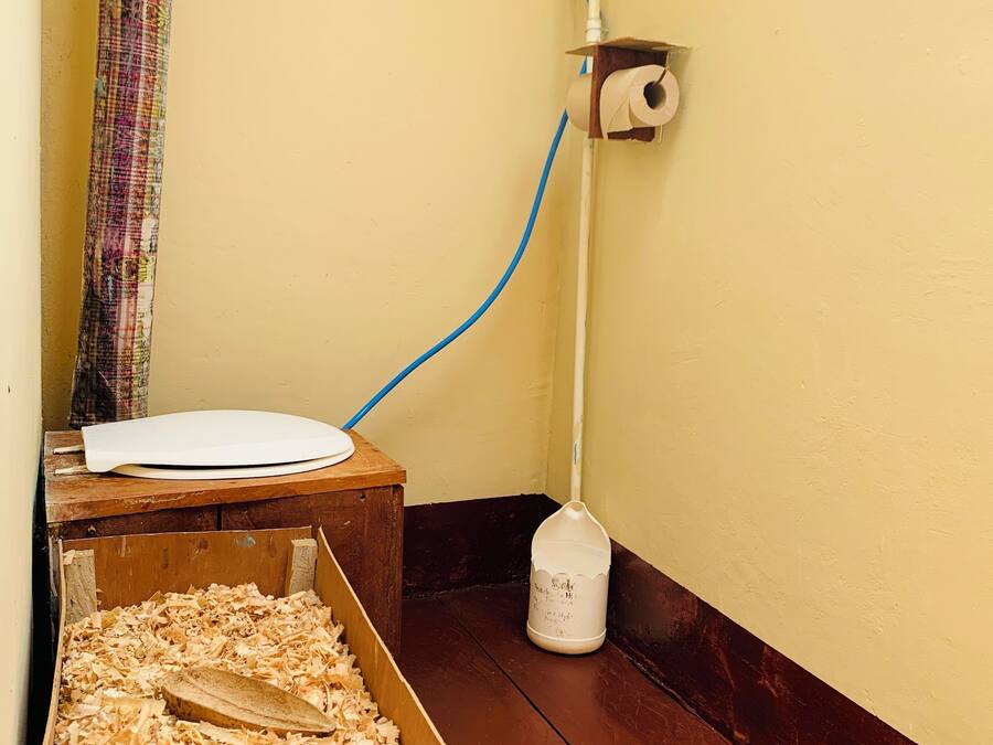 Un racó d'una habitació amb una caixa de fusta plena de virutes, un suport de paper higiènic blanc a la paret i una mànega blava connectada a una font d'aigua.