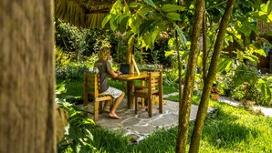 Eine Person sitzt an einem Holztisch mit einem Laptop in einem Garten mit einem reetgedeckten Gebäude.