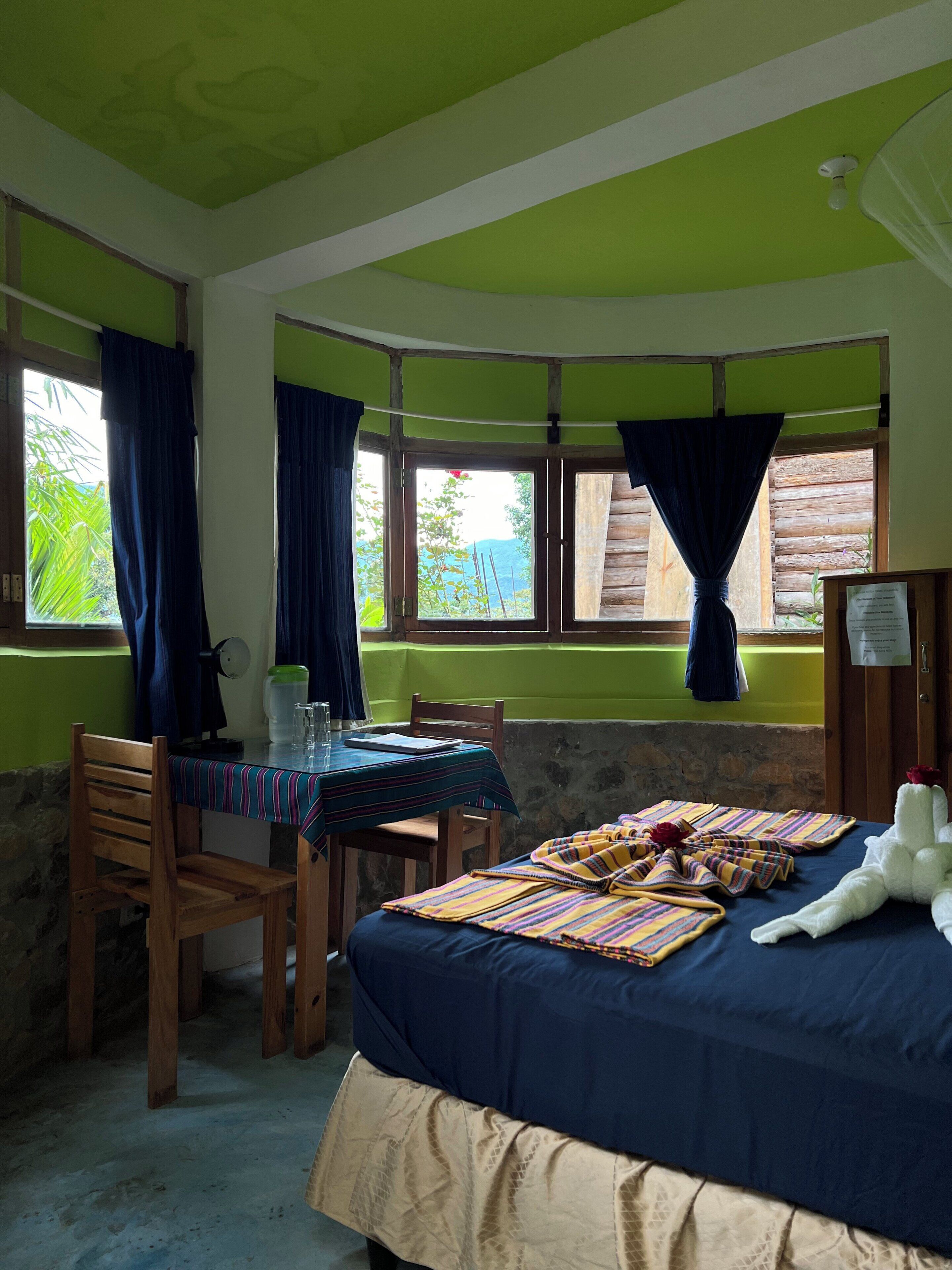 A room with a bed, a table, and chairs. There are windows with curtains, a view of greenery outside, and a ceiling fan.