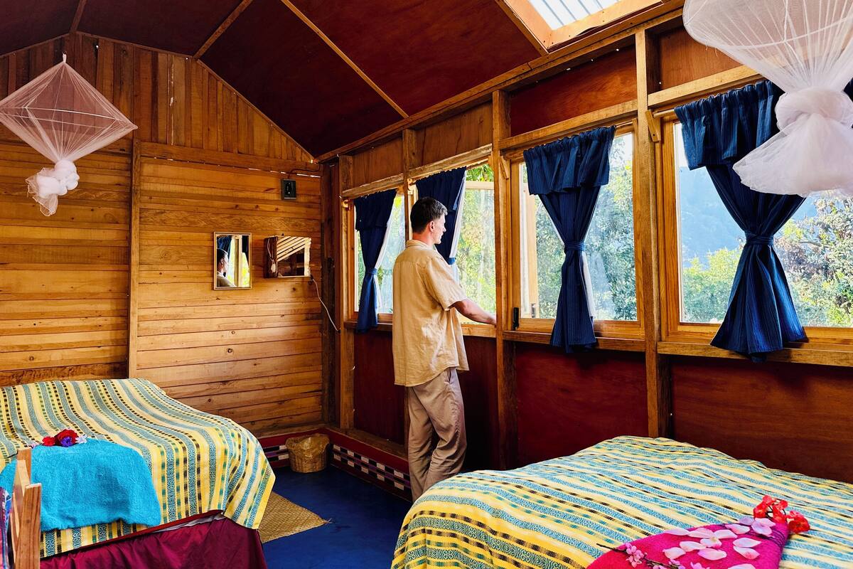 Een houten hutje met twee bedden, een groot raam met gordijnen, een plafondventilator en een kleine spiegel aan de muur.