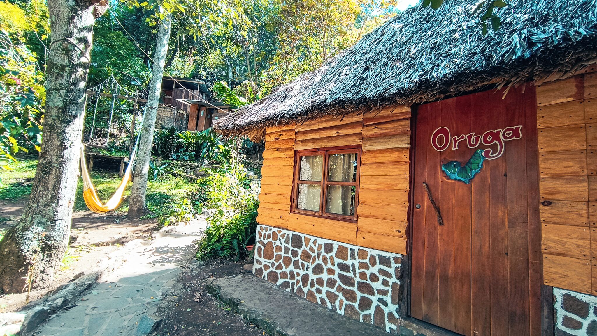 Una cabaña de madera con techo de paja, cimientos de piedra y una hamaca al fondo.